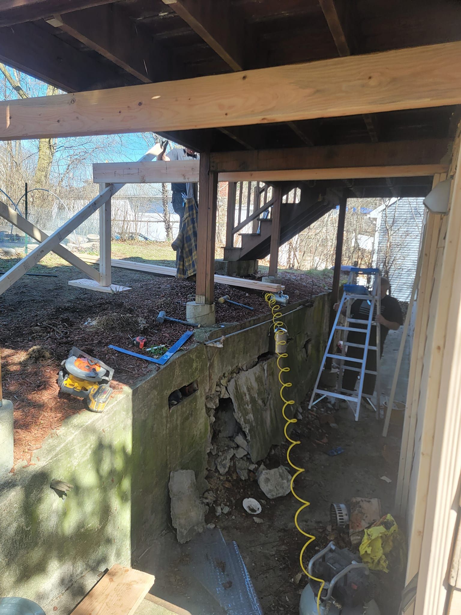 Construction work under a deck. Concrete wall damage. Tools, wood beams, and a ladder are present.