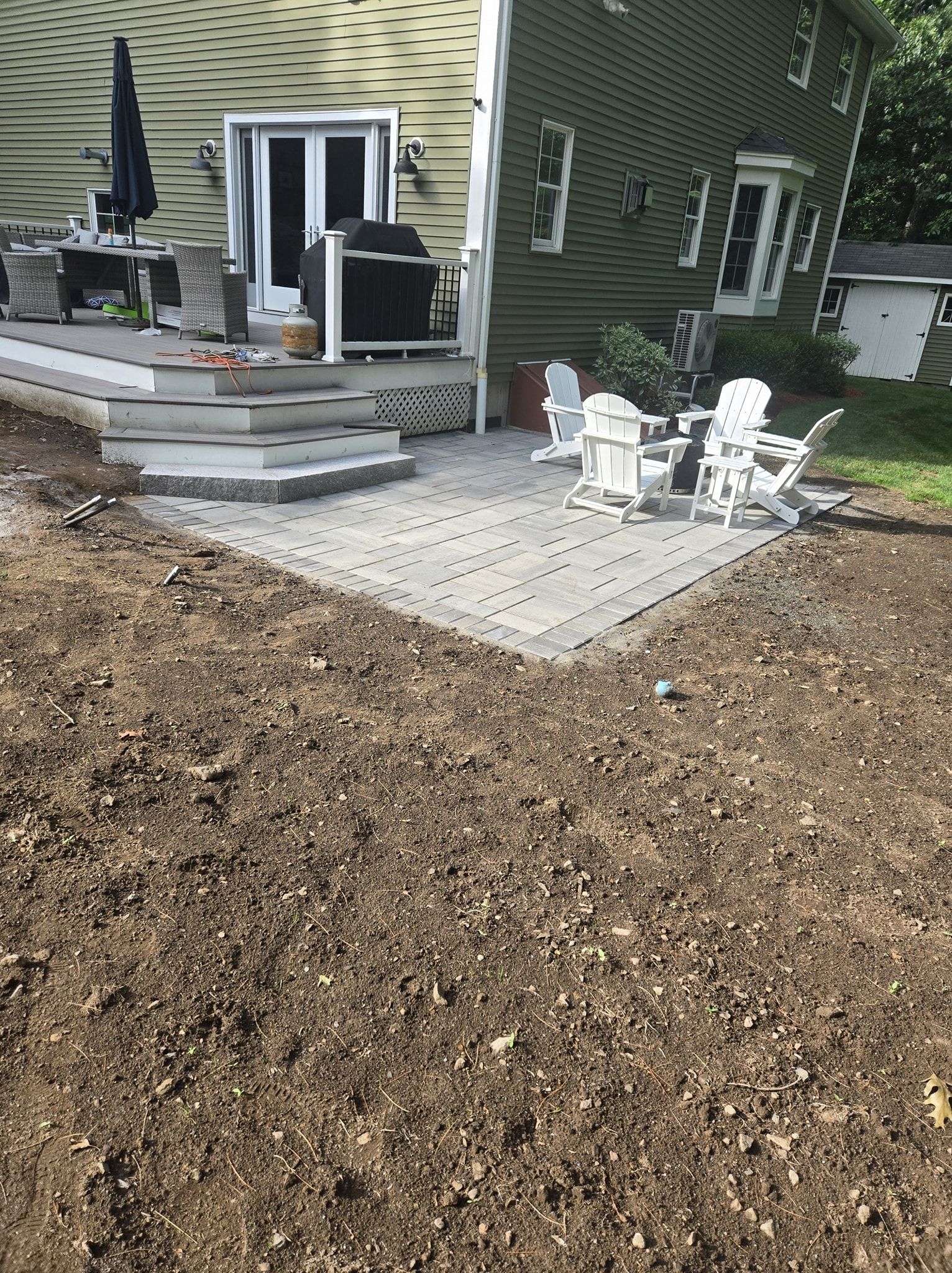 Newly constructed patio and deck with white chairs, next to a green house.