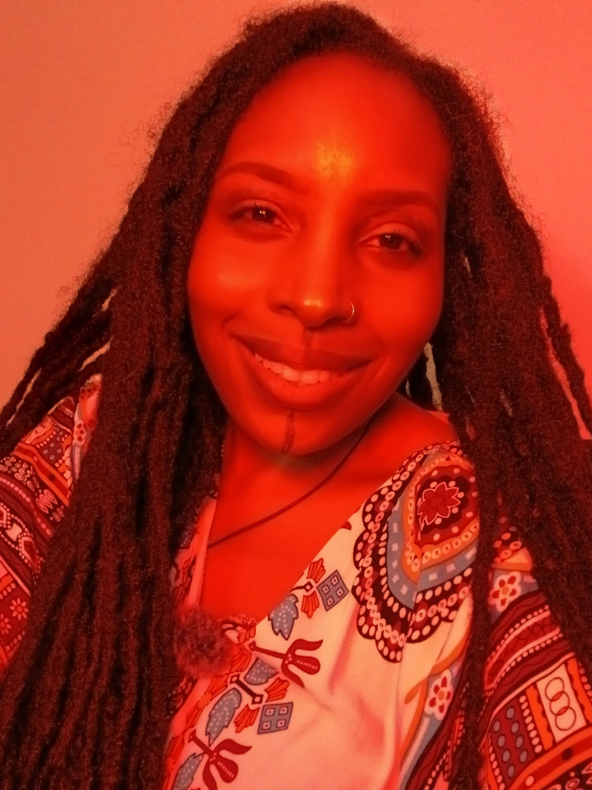 Portrait of a smiling person in red lighting, wearing a patterned top and long braids.
