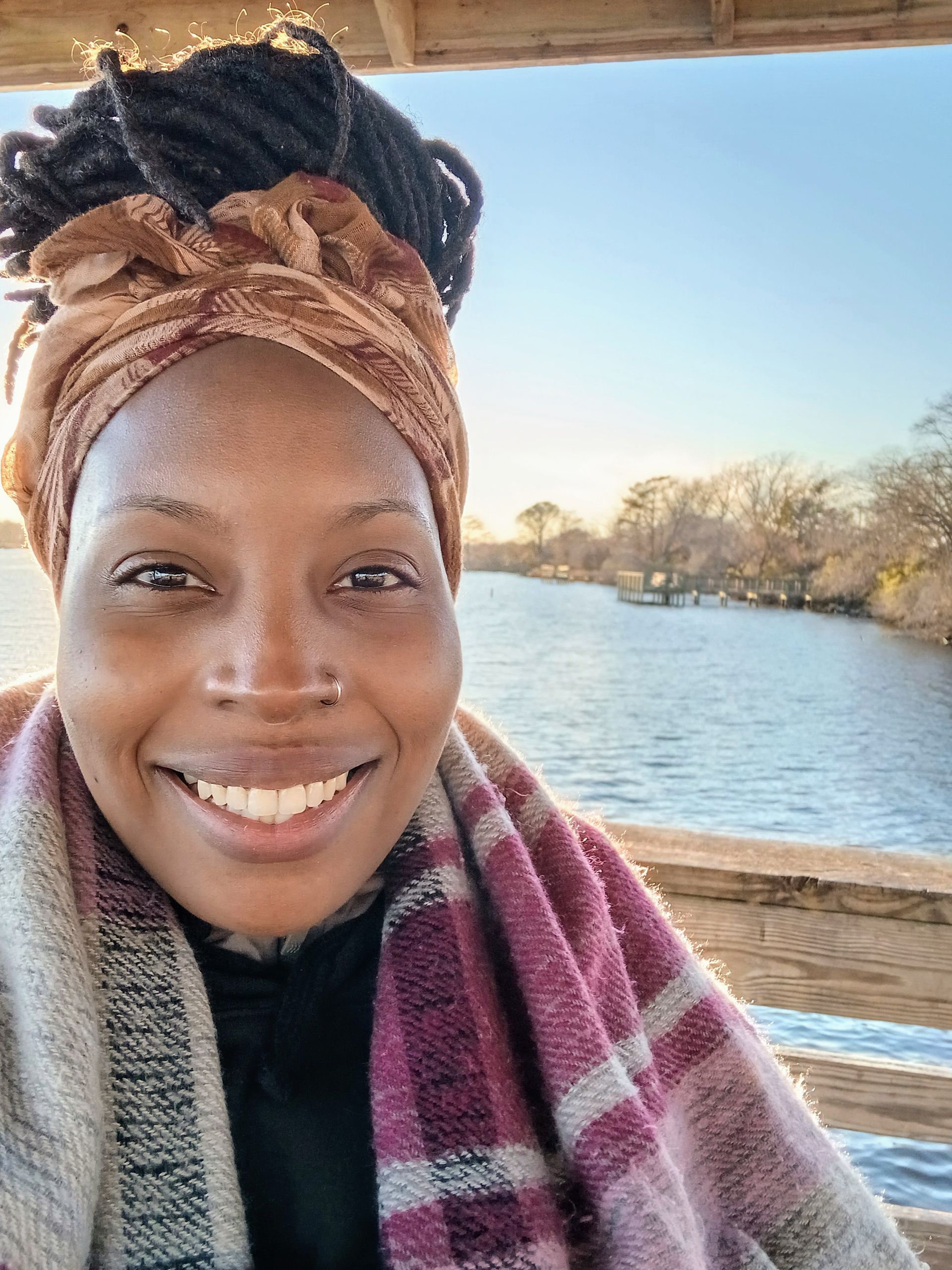 Smiling person in a patterned scarf and headwrap, standing by a riverside with winter trees in the background