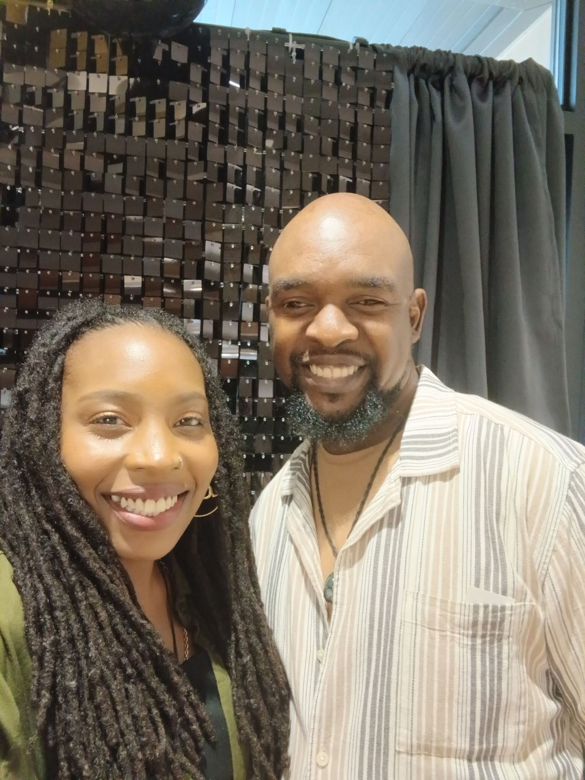 Two smiling people posing indoors in front of a dark curtain and wall of square decor.