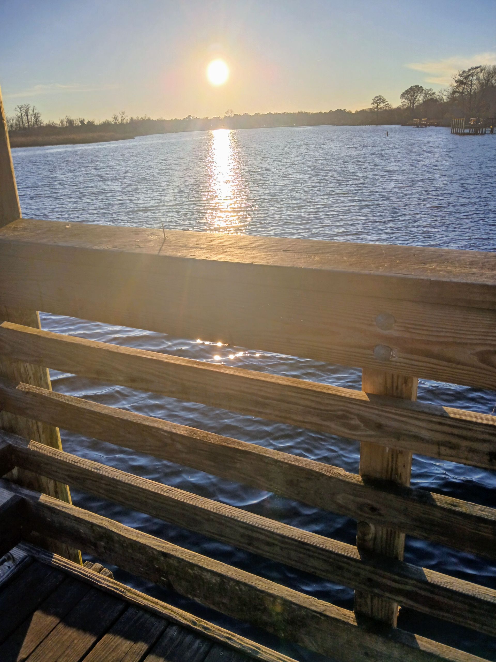 Sunlit lake at sunset viewed from a wooden deck railing, with rippling water and distant trees.