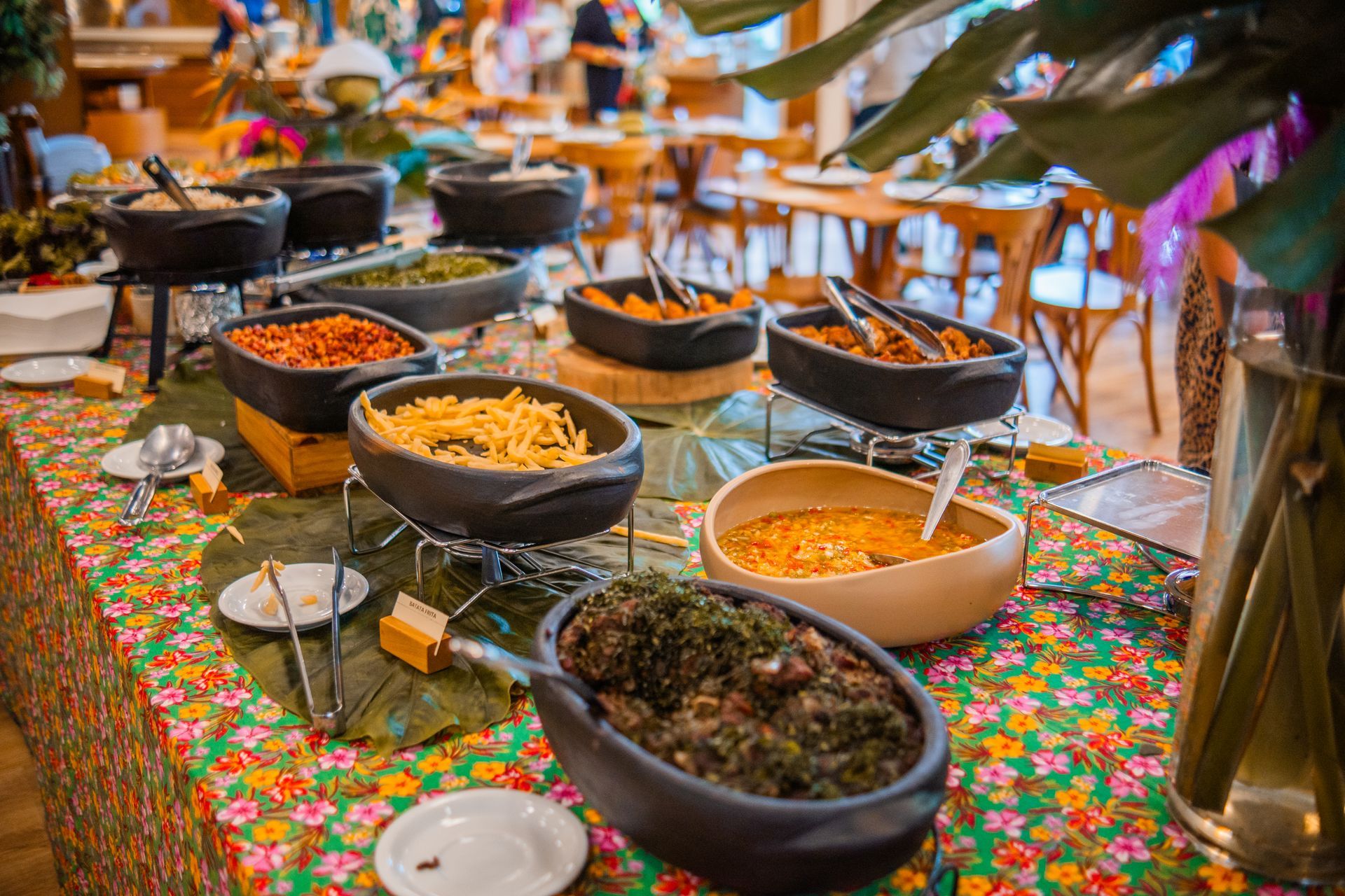 Mesa de buffet com pratos variados em tigelas pretas sobre uma toalha de mesa floral colorida ao ar livre
