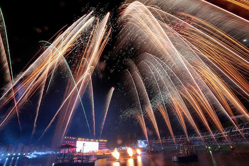 Fogos de artifício explodindo em arcos dourados sobre uma cena noturna à beira-mar.