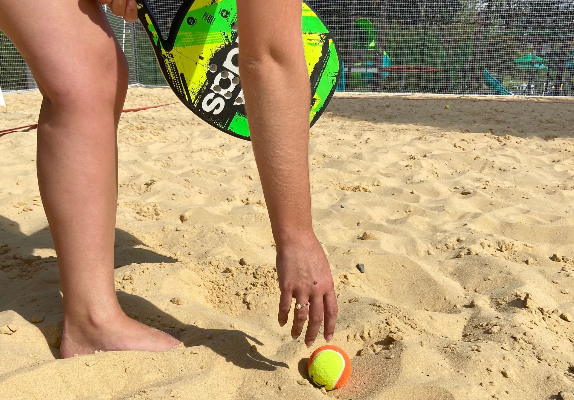 Pessoa estendendo a mão para pegar uma bola de tênis amarela em uma praia arenosa perto de um tabuleiro verde e amarelo.