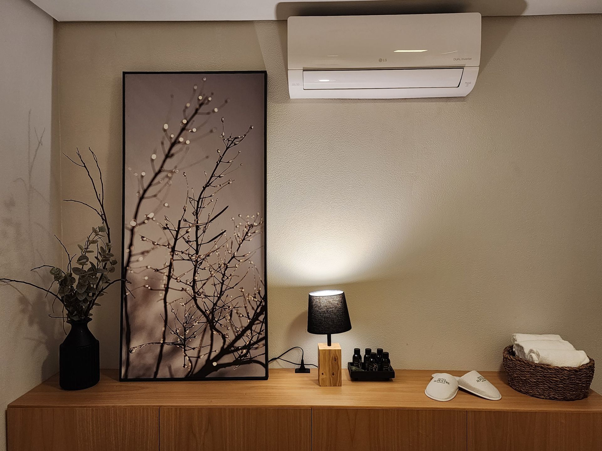 Parede minimalista com estilo de spa, estampa de galhos, ar-condicionado, luminária, toalhas e uma cesta de vime em uma prateleira de madeira.