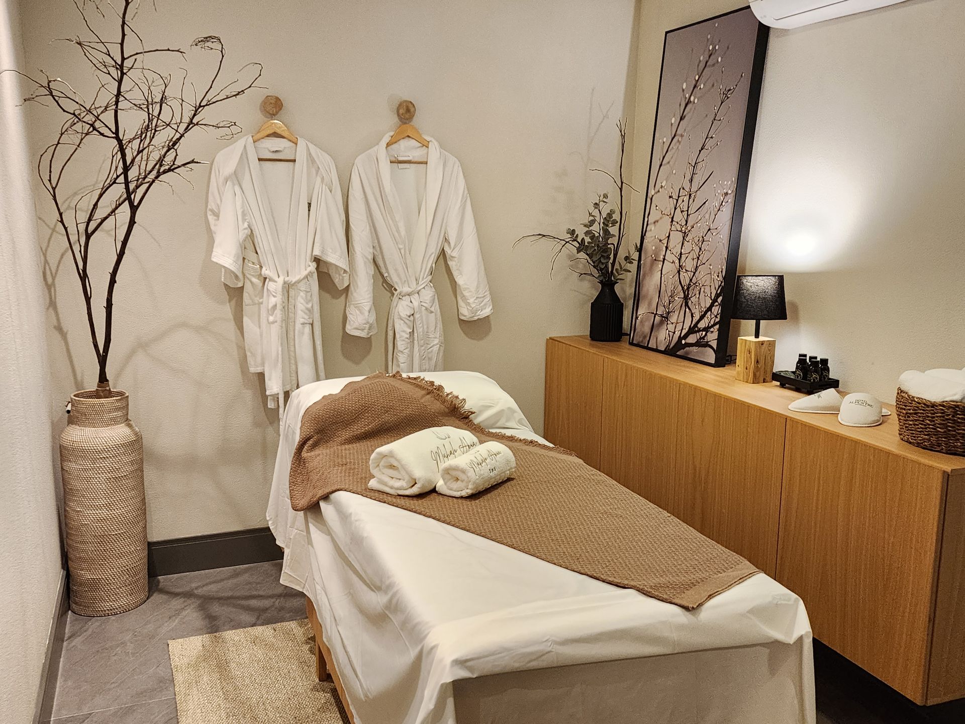 Sala de tratamento de spa com mesa de massagem, roupões brancos, armário de madeira e galhos decorativos.