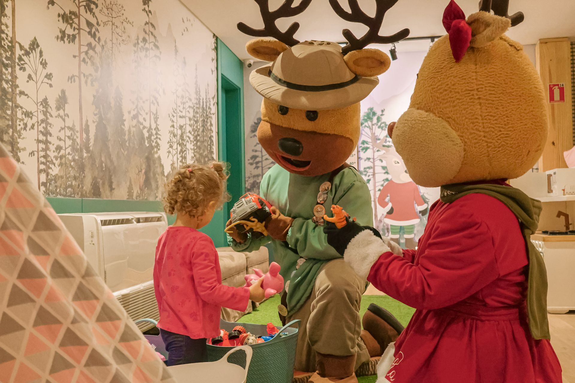 Em um ambiente festivo em um espaço fechado, pessoas posam com mascotes fantasiados de renas e crianças por perto.