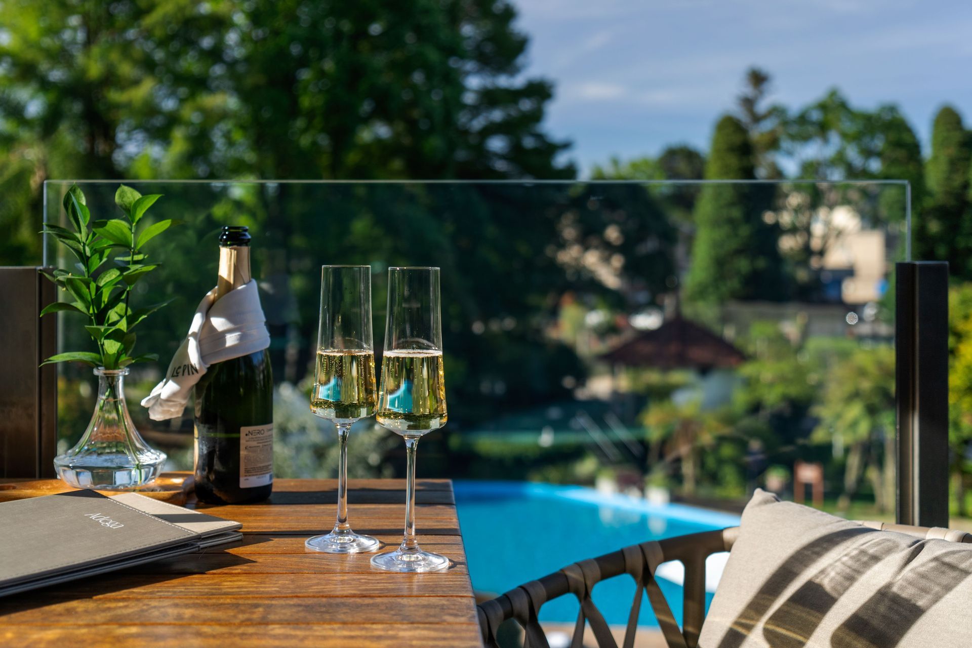 Mesa no deck externo com taças de espumante e jardins e piscina como cenário de fundo.
