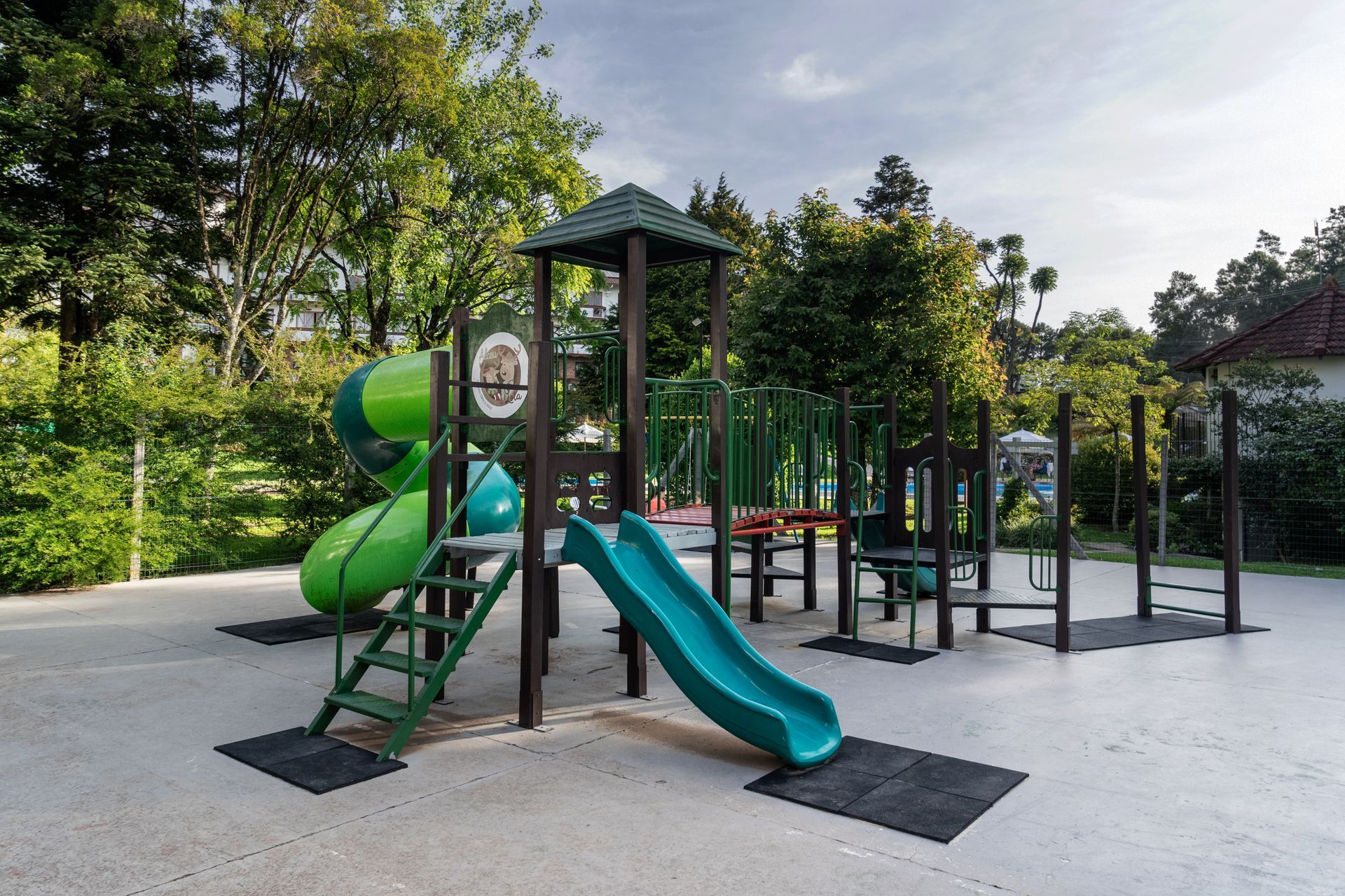 Parque infantil ao ar livre com escorregador espiral verde, escorregador reto azul e estrutura de escalada em piso pavimentado.