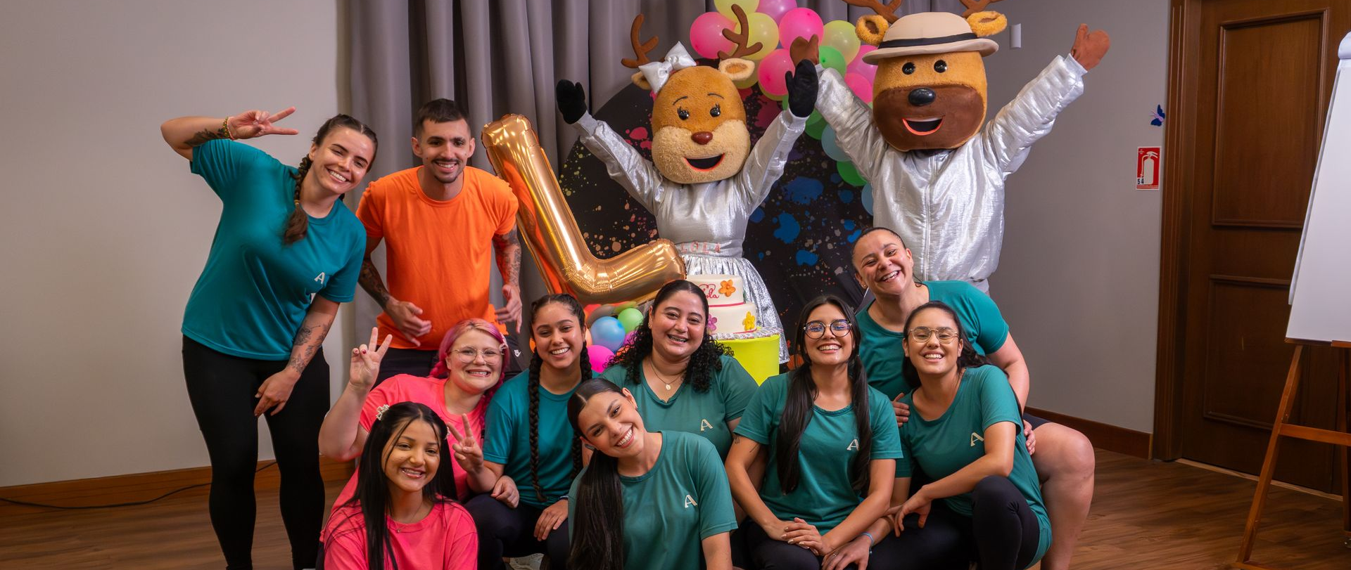 Grupo de pessoas posando em ambiente interno com fantasias coloridas e cabeças de personagens gigantes, sorrindo e acenando.