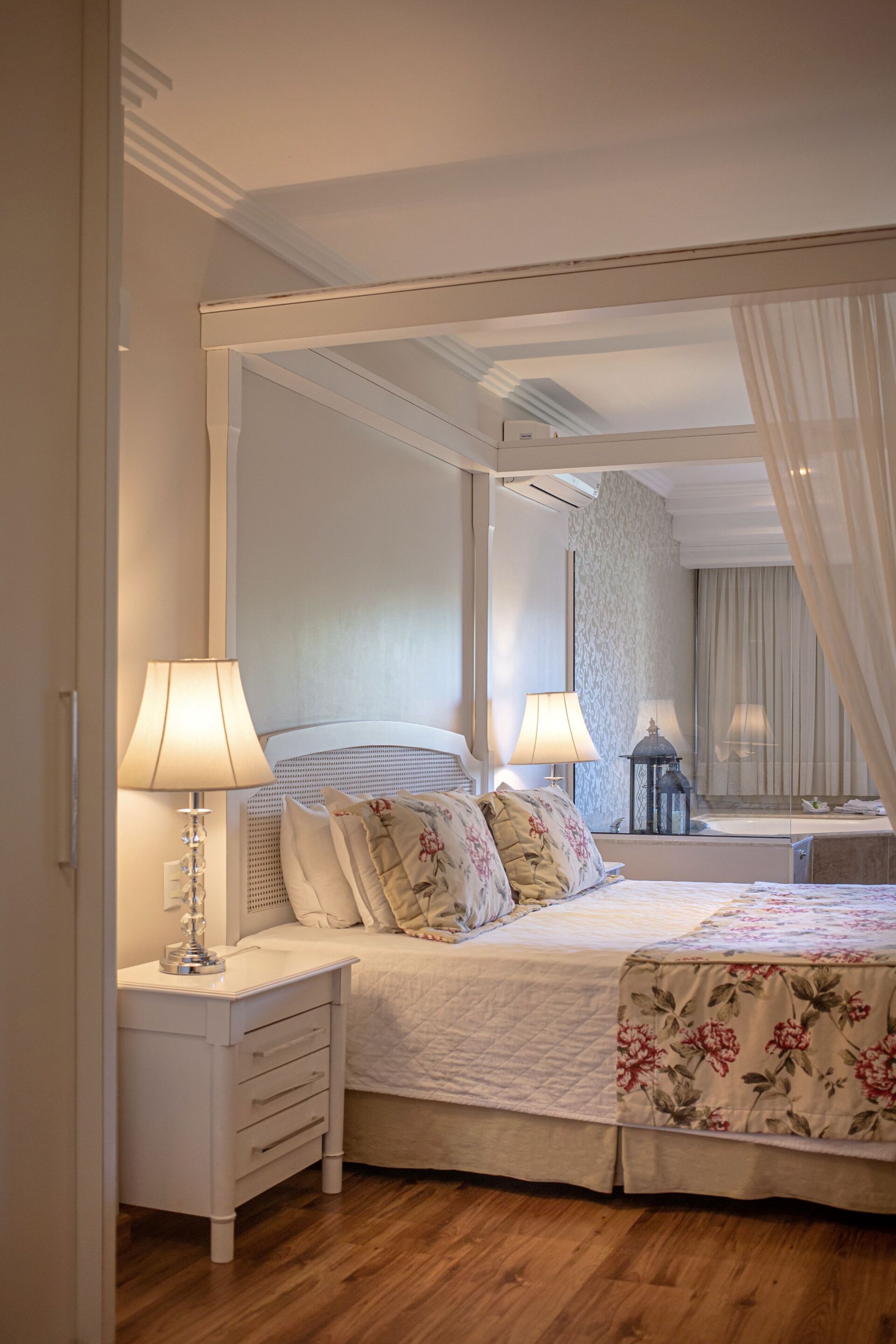 Quarto elegante com dossel branco, roupa de cama floral, abajur e cômoda com espelho.