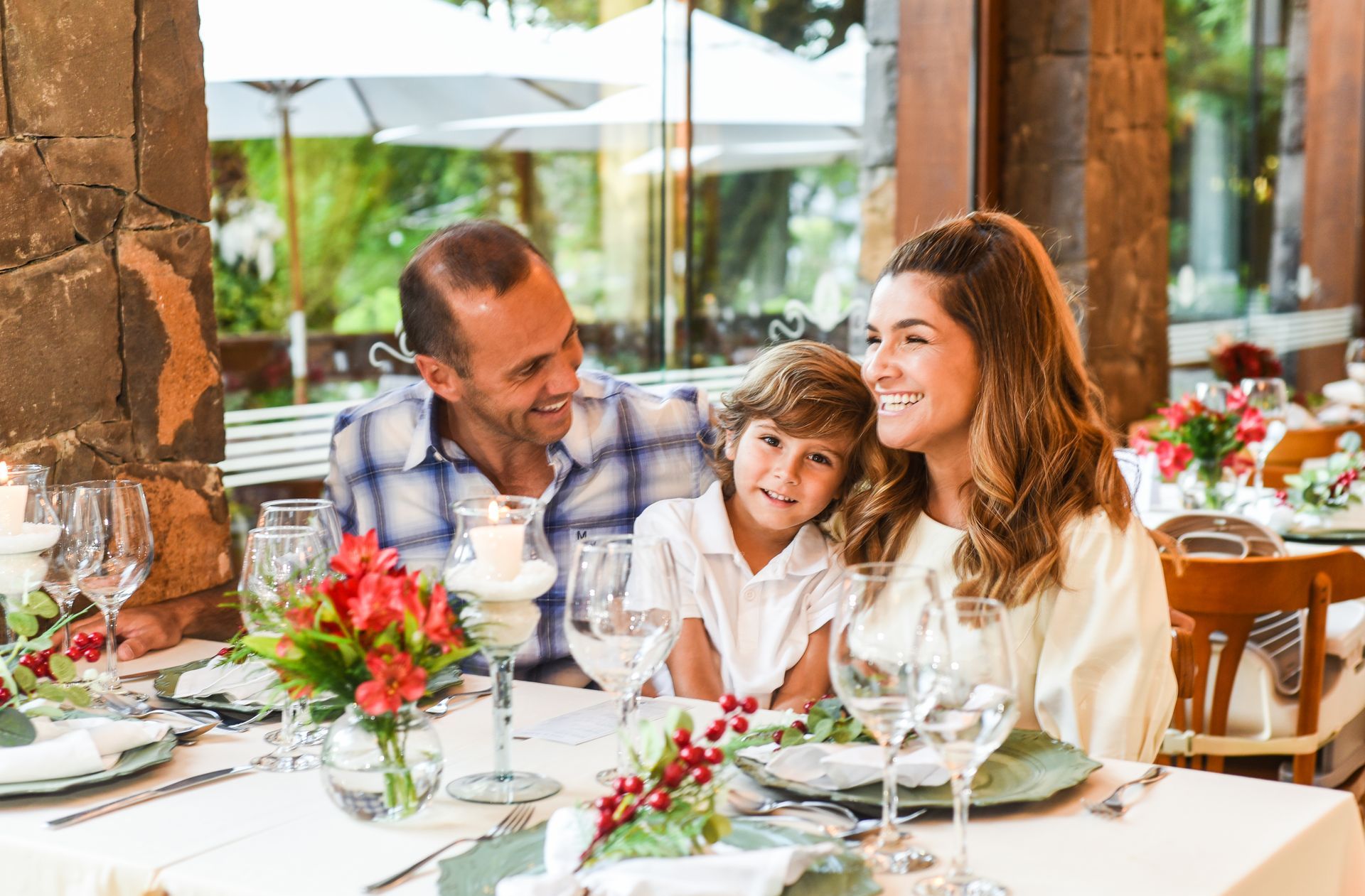 Família reunida para uma refeição num restaurante ao ar livre e bem iluminado, sorrindo à mesa com flores e copos
