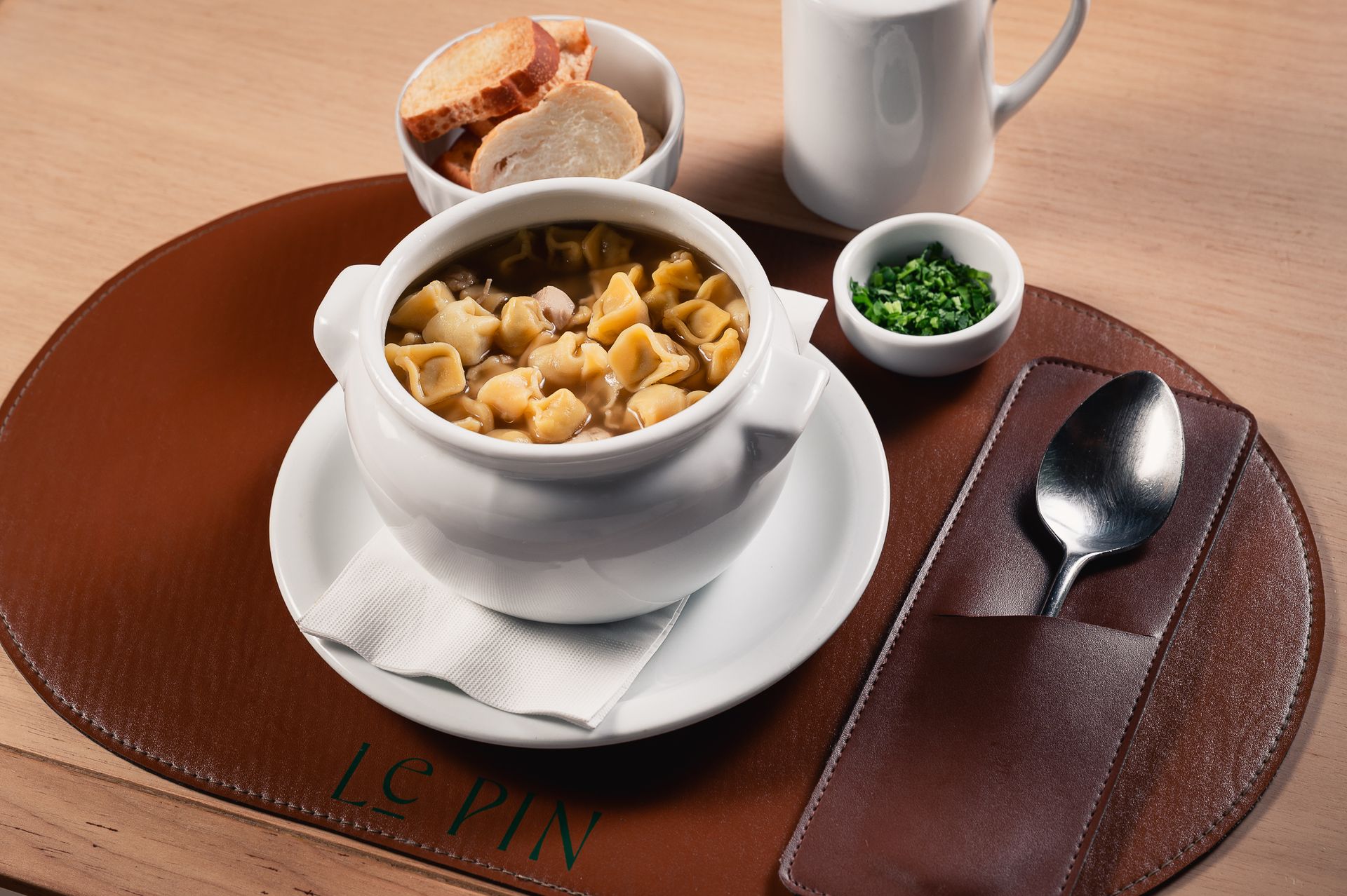 Sopa servida em uma xícara branca sobre um pires, com pão, ervas e uma colher sobre um jogo americano marrom.
