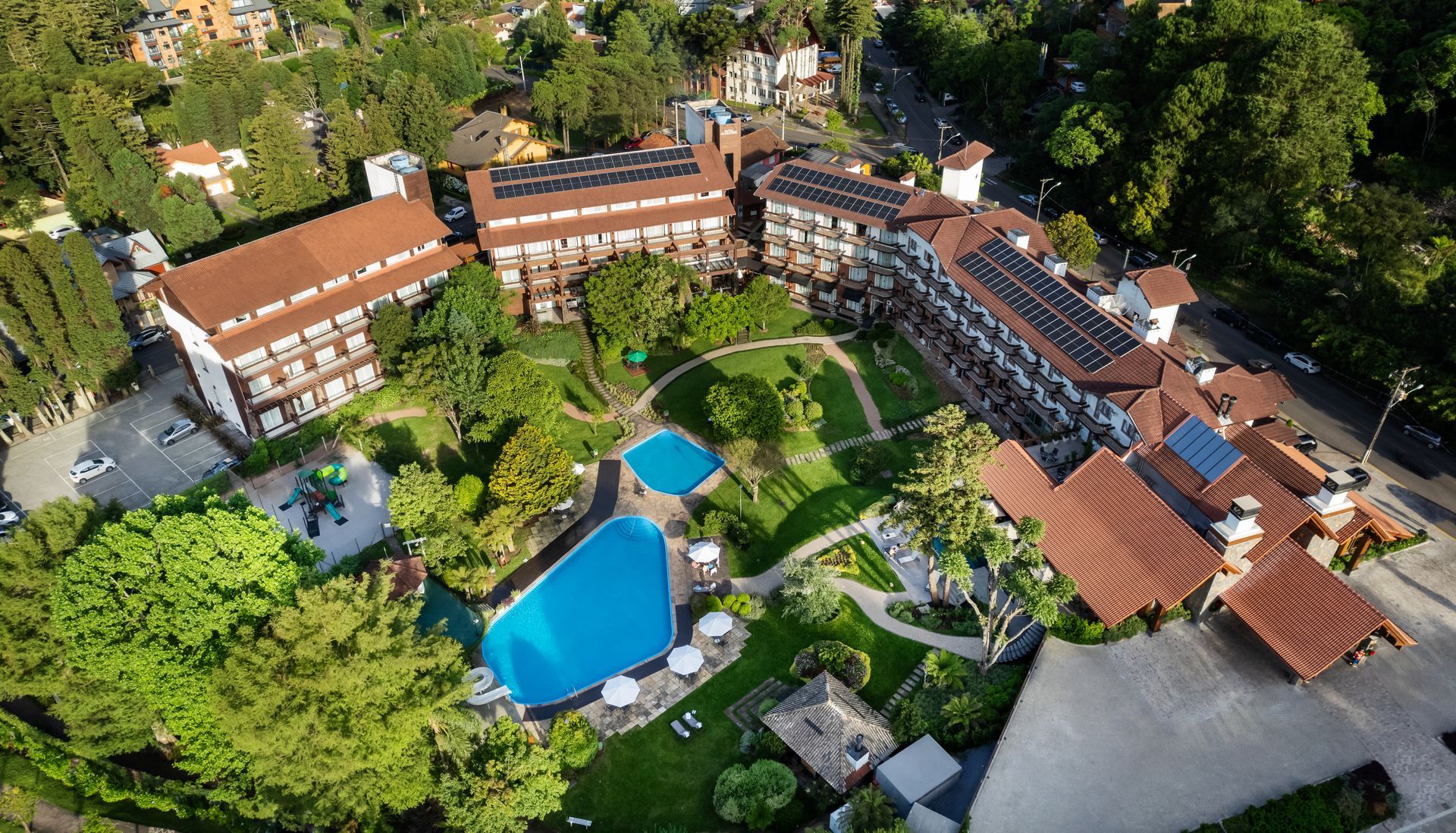Vista aérea de um resort com edifícios de telhados vermelhos, jardins exuberantes e uma piscina azul.