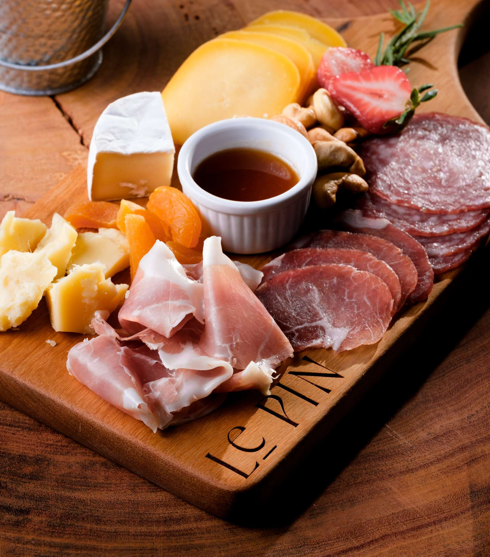 Tábua de charcutaria com queijos, carnes curadas, frutas, nozes e um pequeno recipiente para molho sobre uma mesa de madeira.