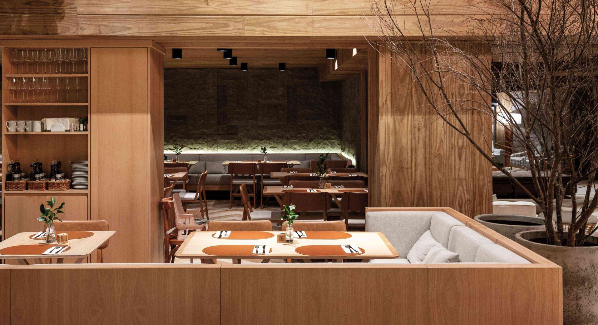 Restaurante aconchegante com painéis de madeira, cabines com almofadas brancas, mesas postas e uma parede de pedra escura ao fundo.