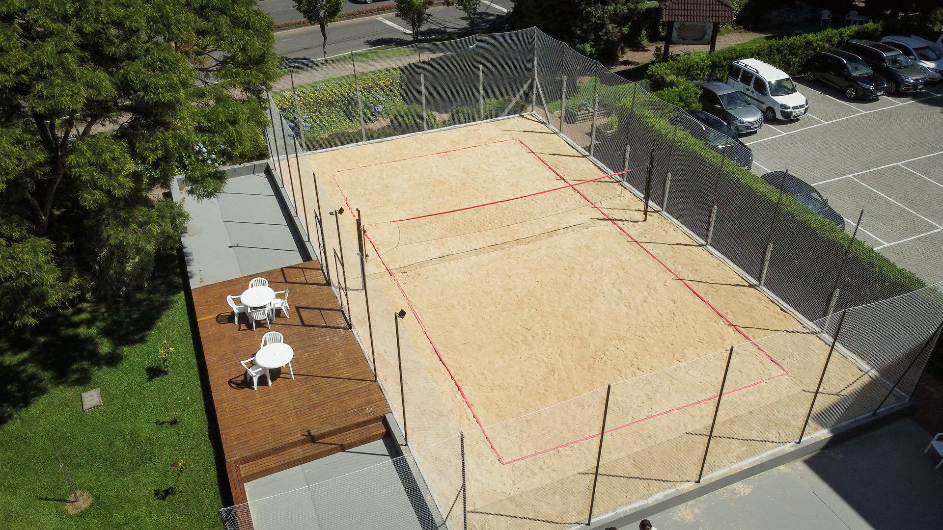 Vista aérea de uma quadra de vôlei de areia ao ar livre, ao lado de um deck com mesas e cadeiras em um estacionamento.