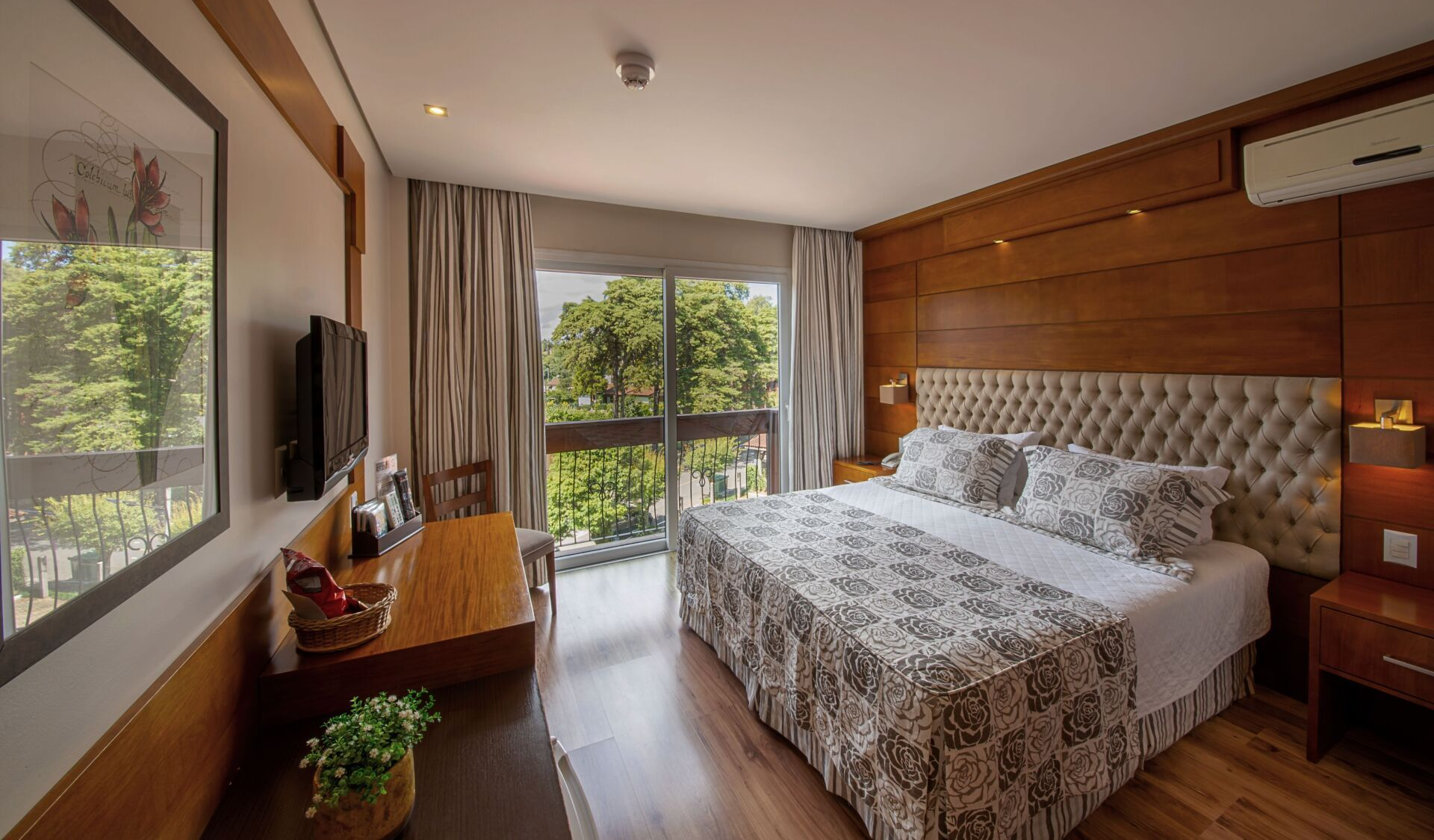Quarto moderno com cama grande, parede revestida de madeira, TV e portas de varanda com vista para a vegetação.