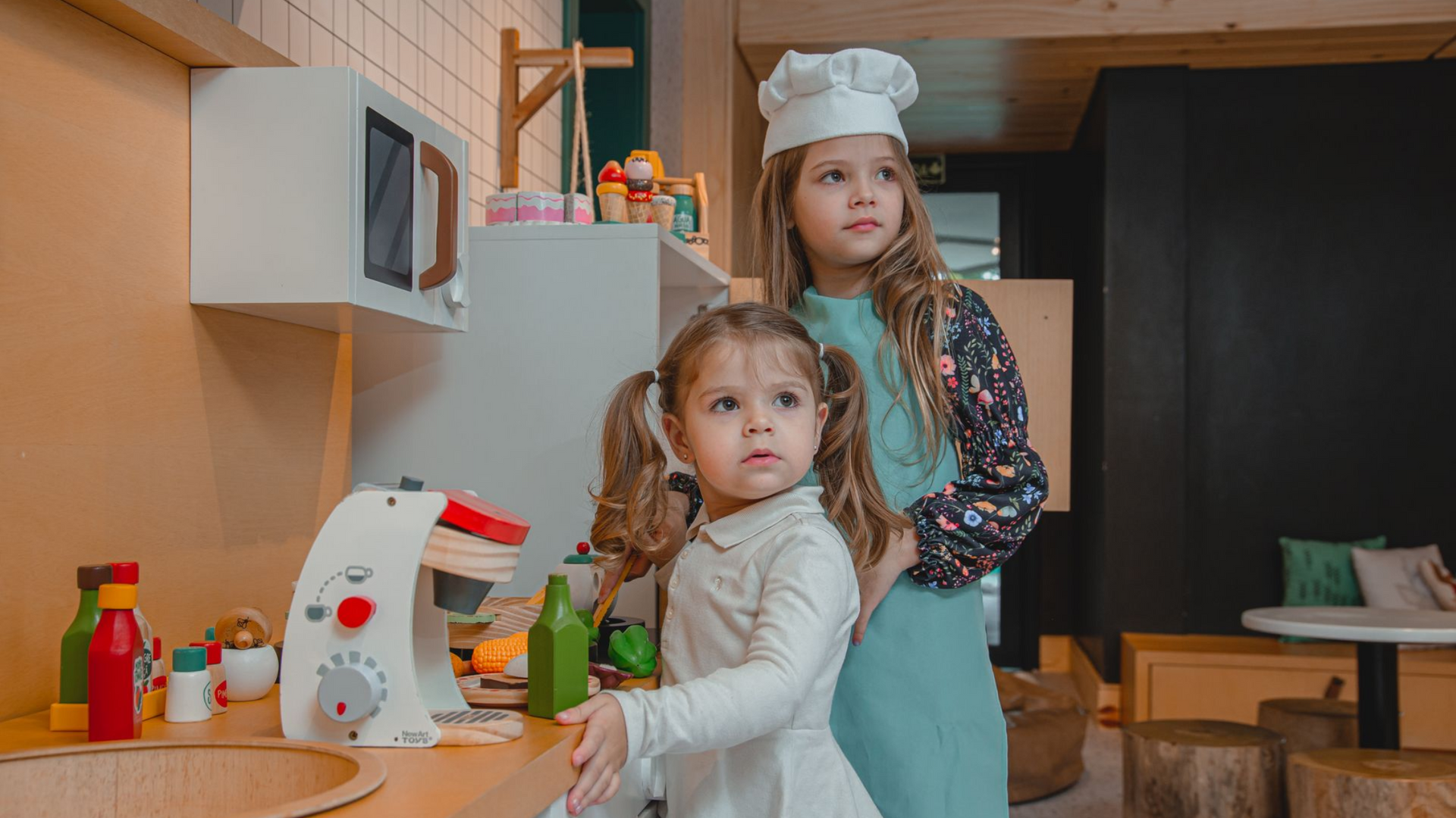 Duas crianças de avental e chapéu de chef estão em pé em uma bancada de cozinha com comidinhas de brinquedo e outros brinquedos.