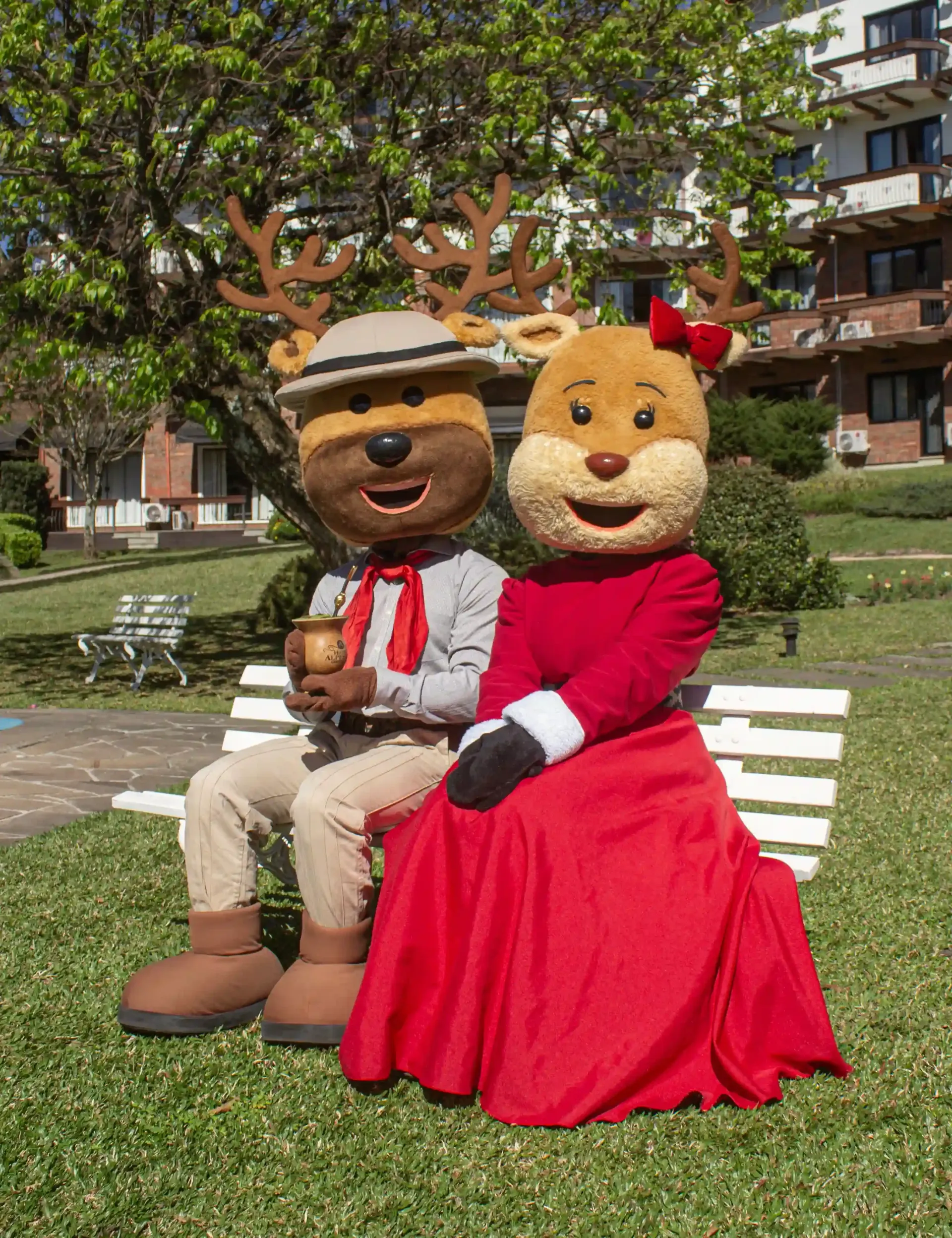 Dois mascotes em trajes festivos sentados em um banco branco em um parque gramado.