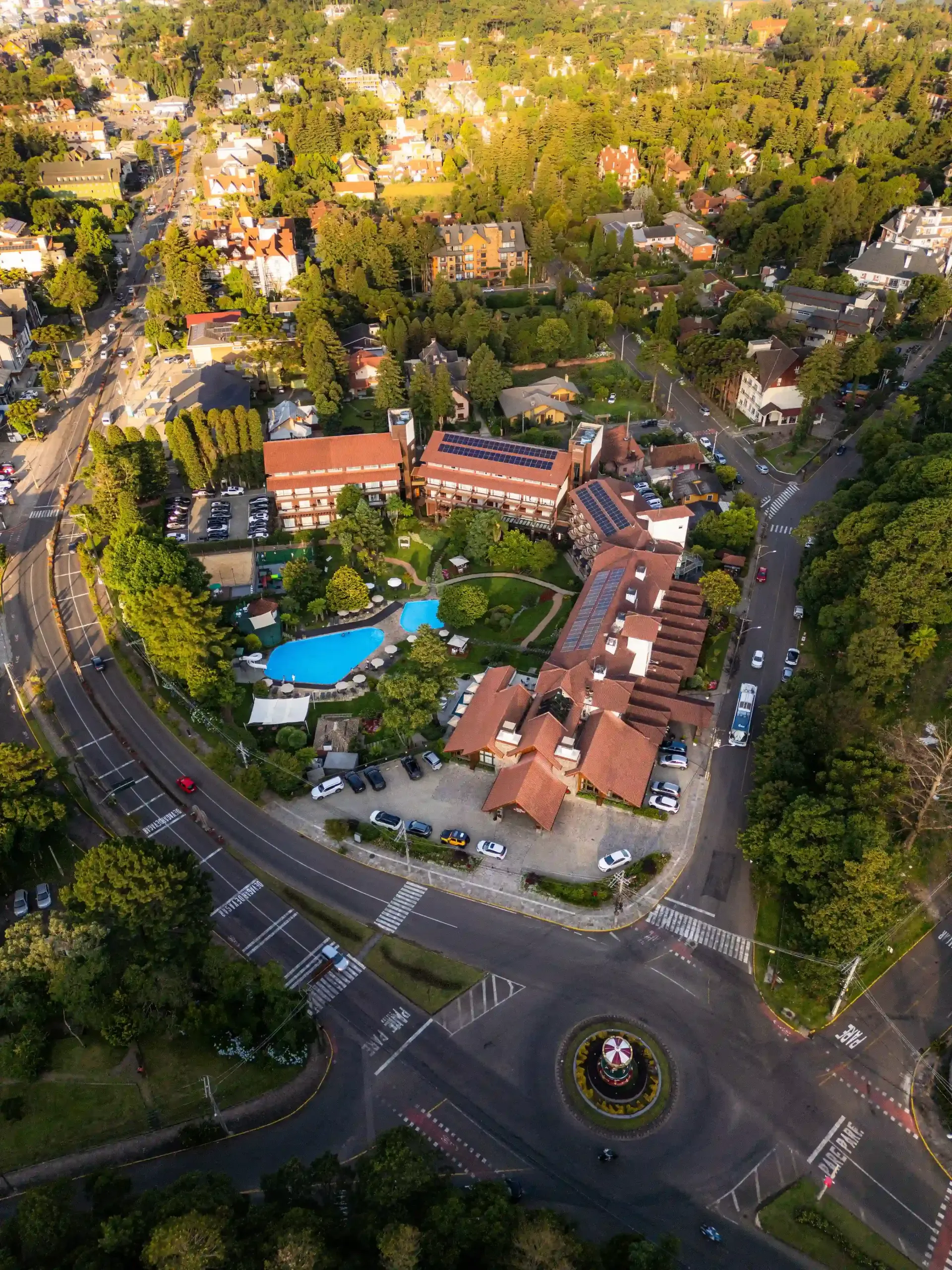 Vista aérea de um resort com edifícios de telhados vermelhos, uma piscina azul e uma rotunda rodeada de árvores.