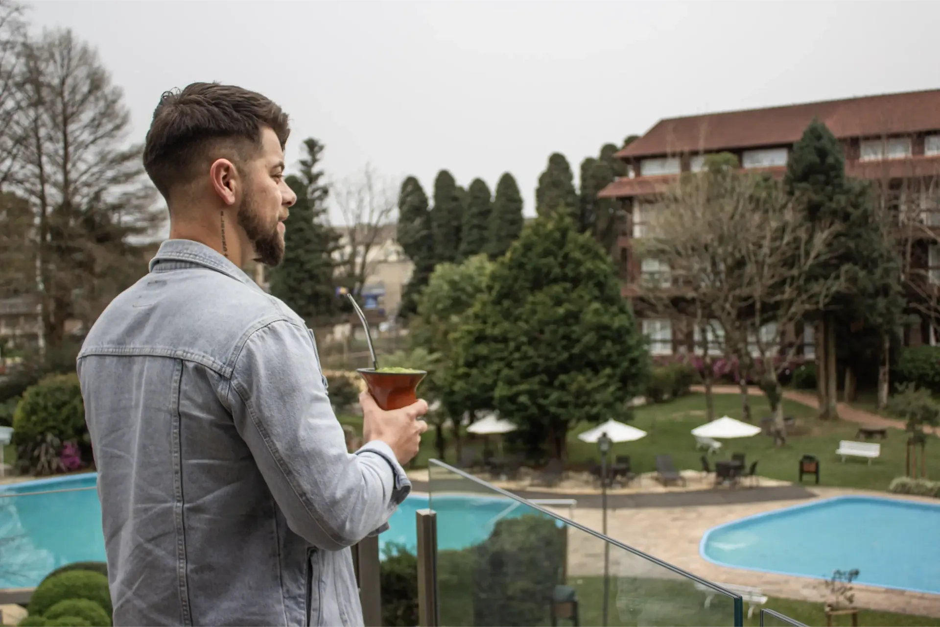 Homem segurando uma cuia de chimarrão em uma varanda com vista para a piscina e os jardins do resort.