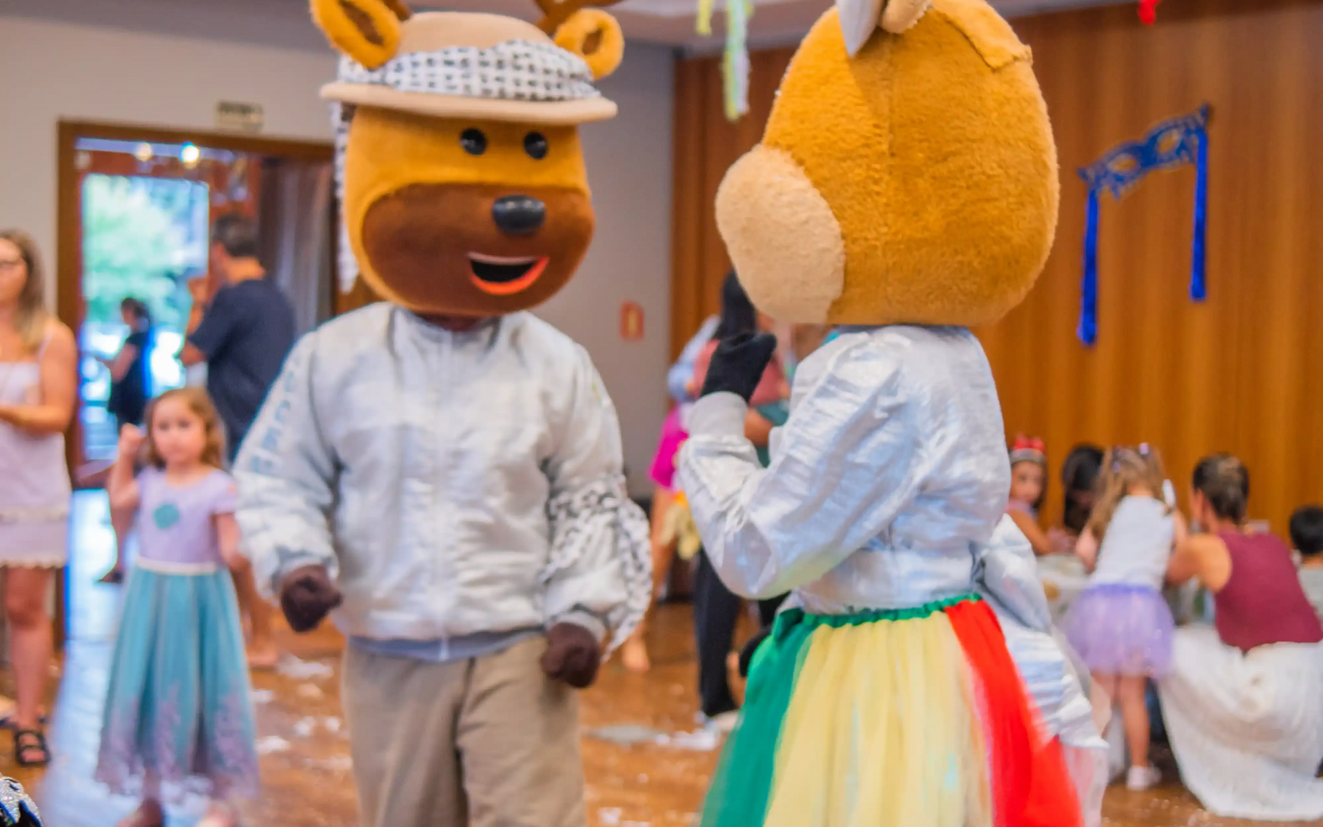 Pessoa sorridente sentada entre dois mascotes em tamanho real em um ambiente interno, um vermelho e outro verde-azulado.