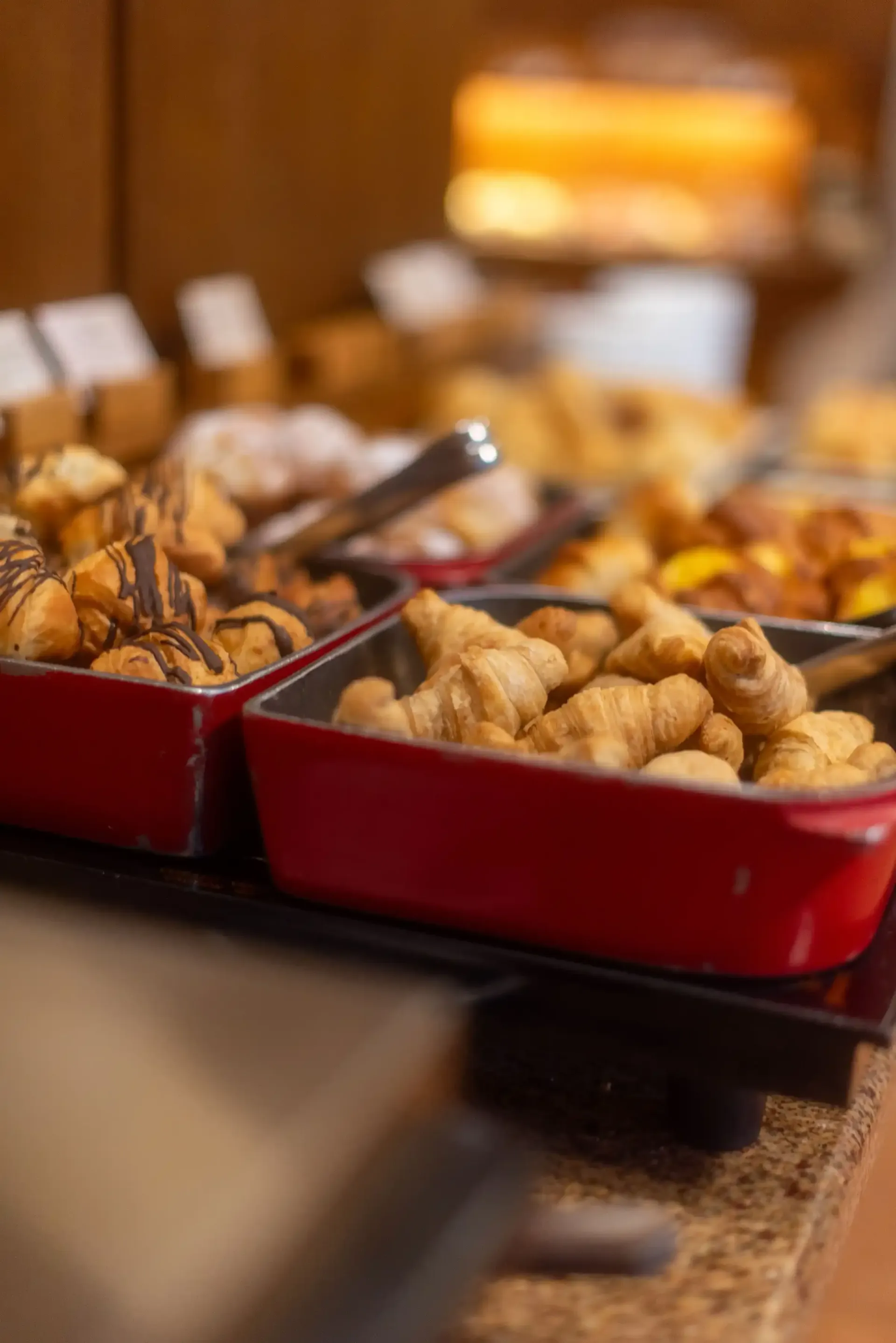 Bandejas vermelhas com diversos petiscos fritos em um balcão de buffet, com pegadores, iluminação interna aconchegante.