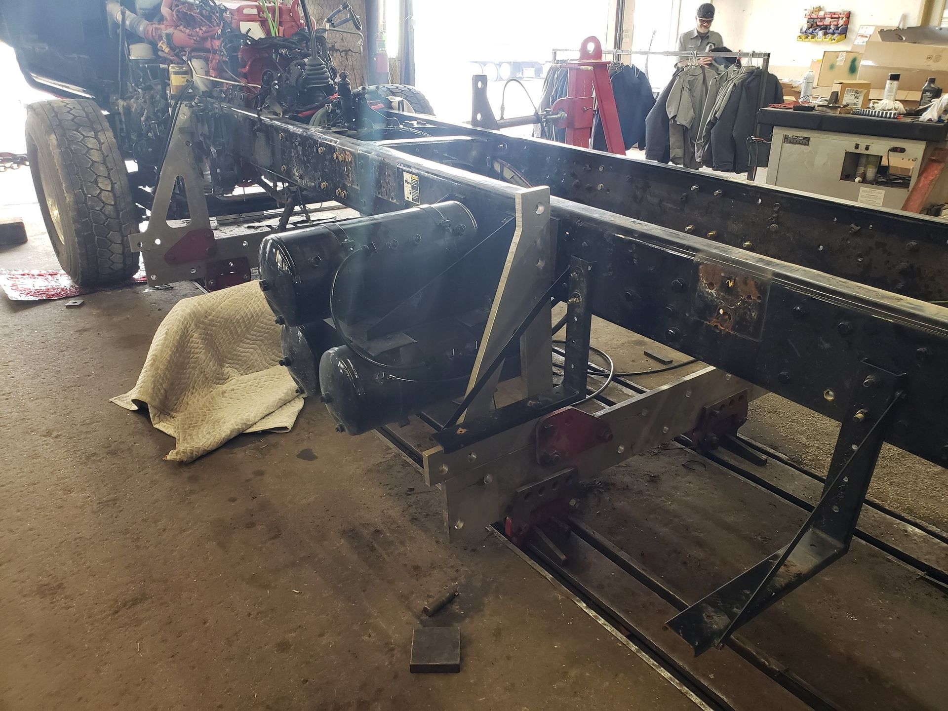Truck frame on stands in a workshop. Black frame, silver metal supports. A person is visible in the background.