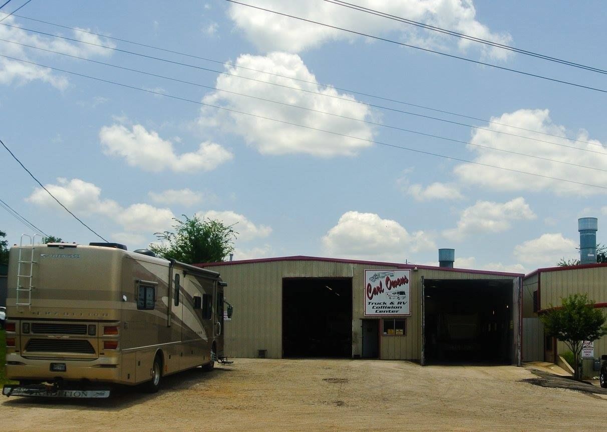 RV parked in front of an auto repair shop with open bays under a cloudy sky.