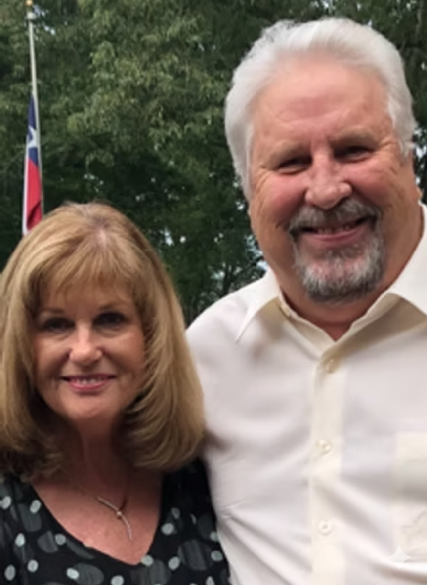 Smiling man and woman pose for photo outdoors; he has white hair and beard, she has blonde hair.