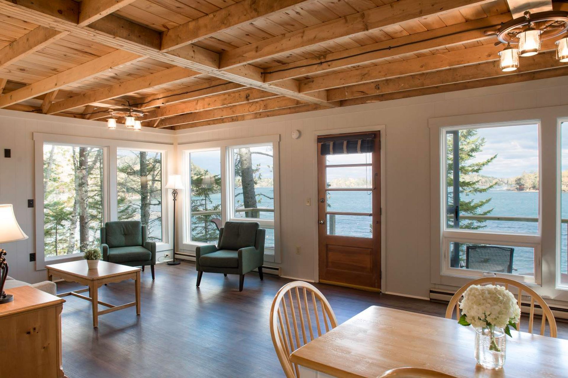 Bright interior living space with lake view, wood ceiling, large windows, two armchairs, and dining table.