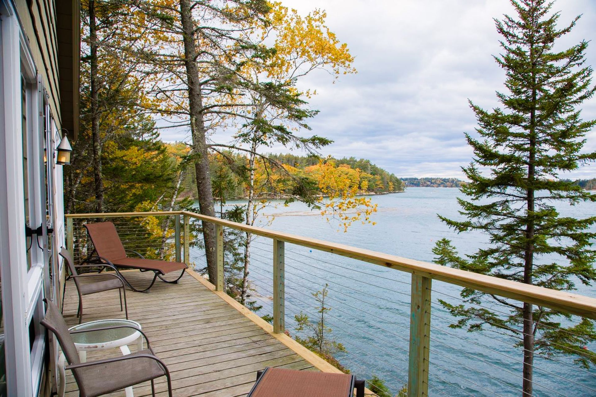 Wooden deck with lounge chairs overlooks a lake surrounded by trees with fall colors under a cloudy sky.