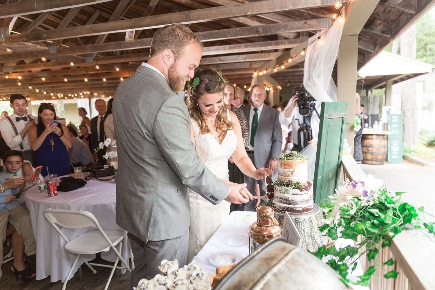 Bride and groom cutting wedding cake at outdoor reception, people watch.