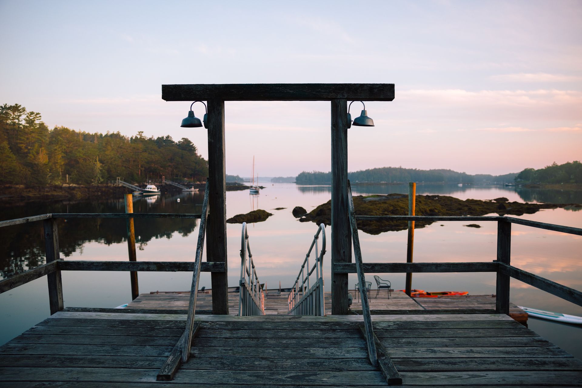 A wooden dock frames a calm lake at sunrise, with boats and distant hills reflected in the water.