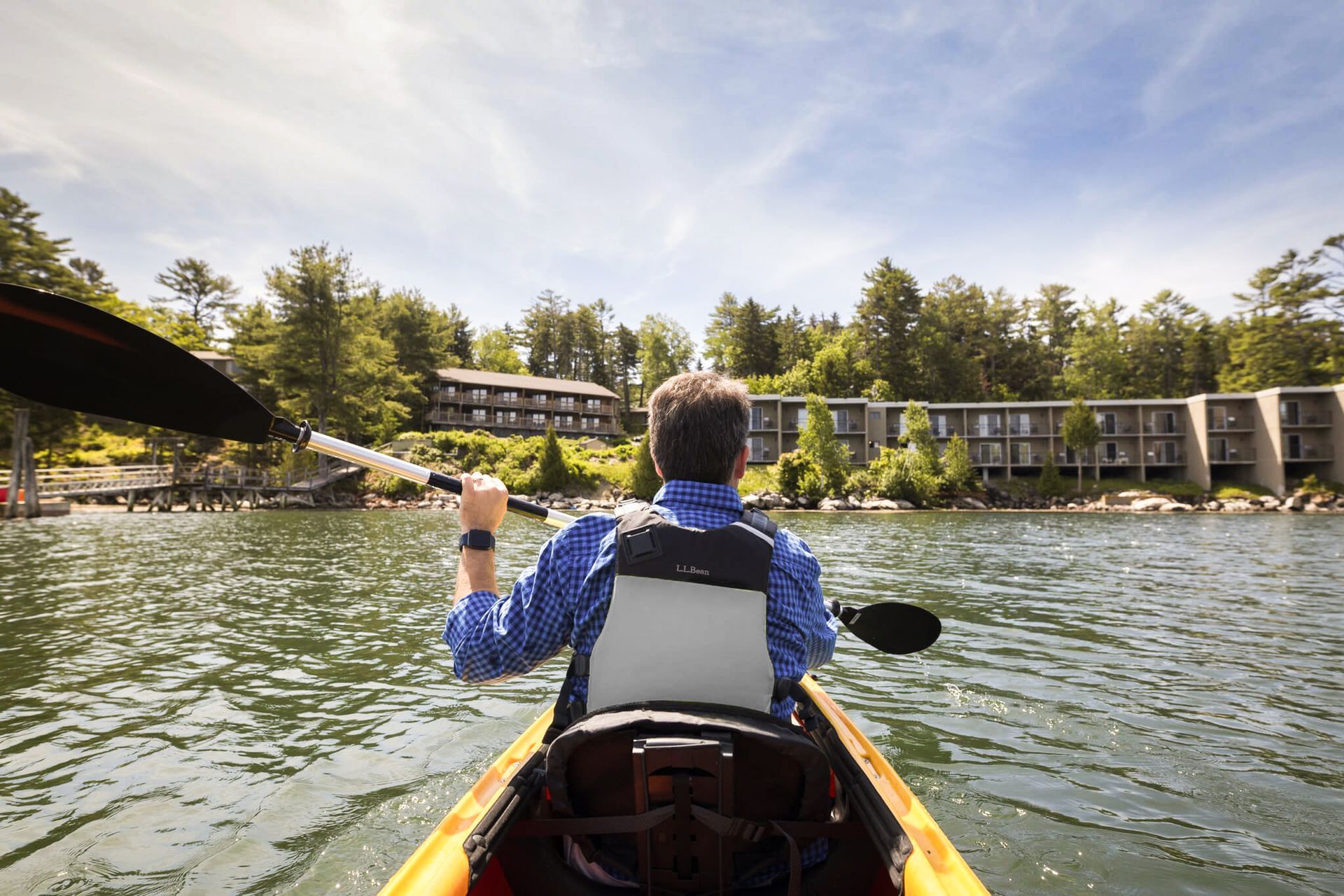 Person kayaking on water, approaching waterfront buildings on a sunny day.