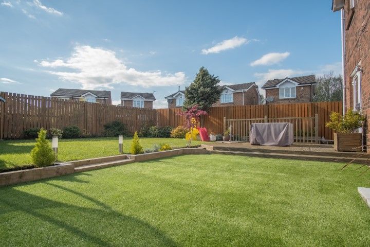A sunny, well-maintained backyard with a two-level lawn, wooden fencing, and a raised wooden deck with a covered table.