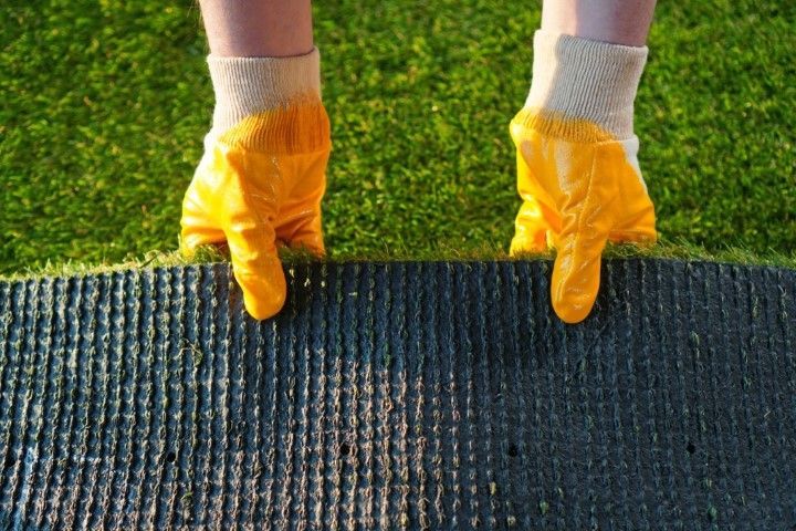 Hands in bright yellow rubber work gloves lift a section of artificial grass to reveal the black backing underneath.