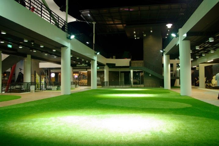 A modern outdoor commercial area at night, featuring a green artificial grass courtyard illuminated by overhead lighting.