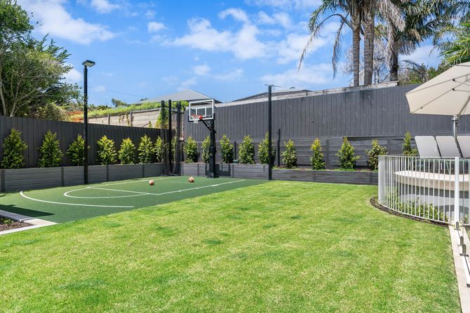 A backyard scene with a green turf basketball court, a hoop, and a grassy lawn enclosed by a tall fence and hedges.