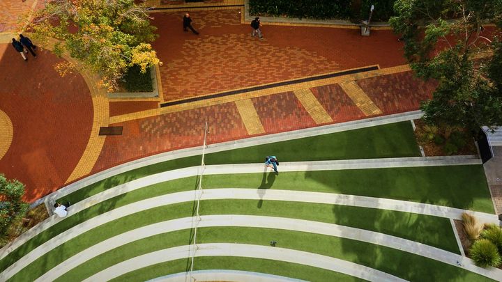 High-angle shot of a park with curved, tiered grassy seating, paved walkways, and people walking through the space.
