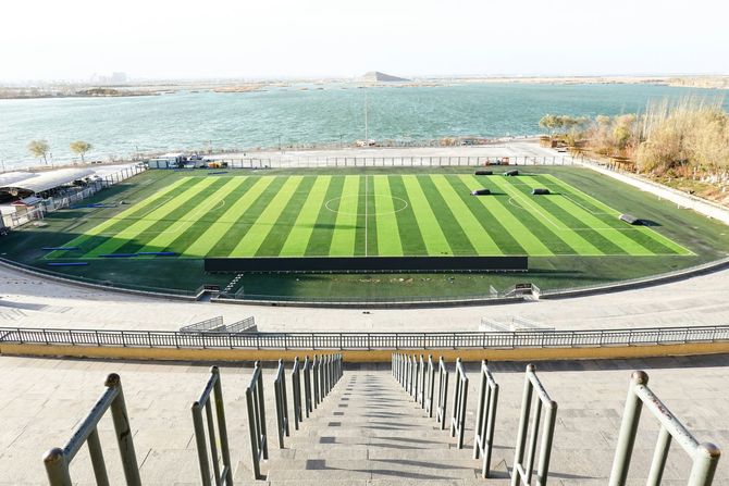An empty soccer field with alternating light and dark green stripes, situated next to a large body of water.