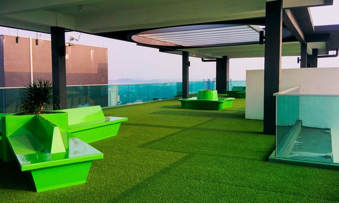 A rooftop terrace with bright green geometric benches on artificial turf, featuring a glass railing overlooking a city.