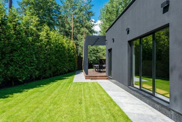 A modern dark gray house with large glass windows borders a manicured green lawn and a stone path leading to a patio.