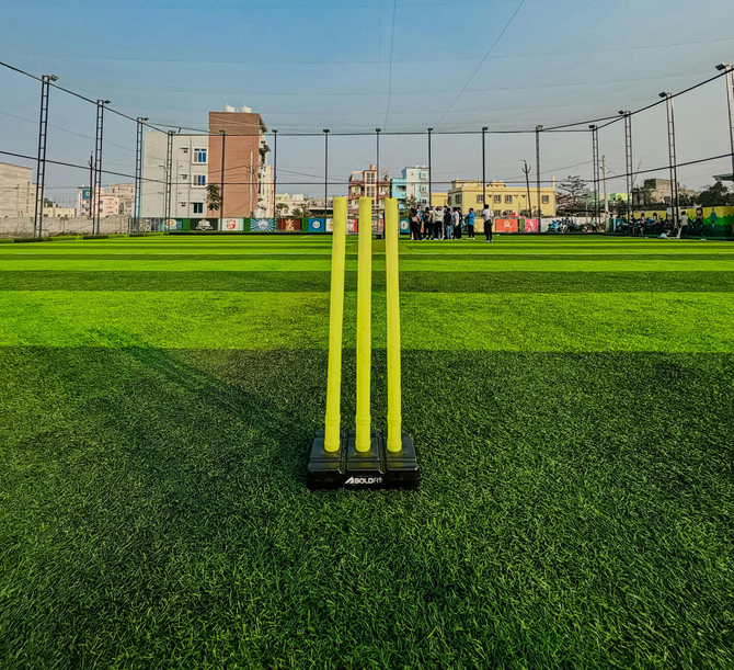 Yellow cricket stumps stand on a green artificial turf field at an outdoor facility with distant buildings.