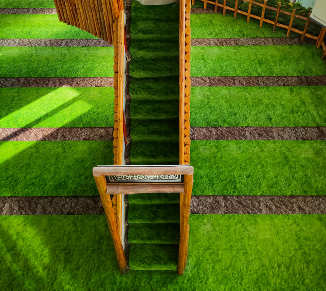 A top-down view of a wooden staircase covered in green artificial grass, set amidst a lawn with horizontal brown paths.