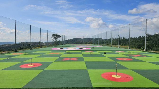 A checkered green and lime turf field for golf practice, featuring circular target pads and surrounded by safety netting.