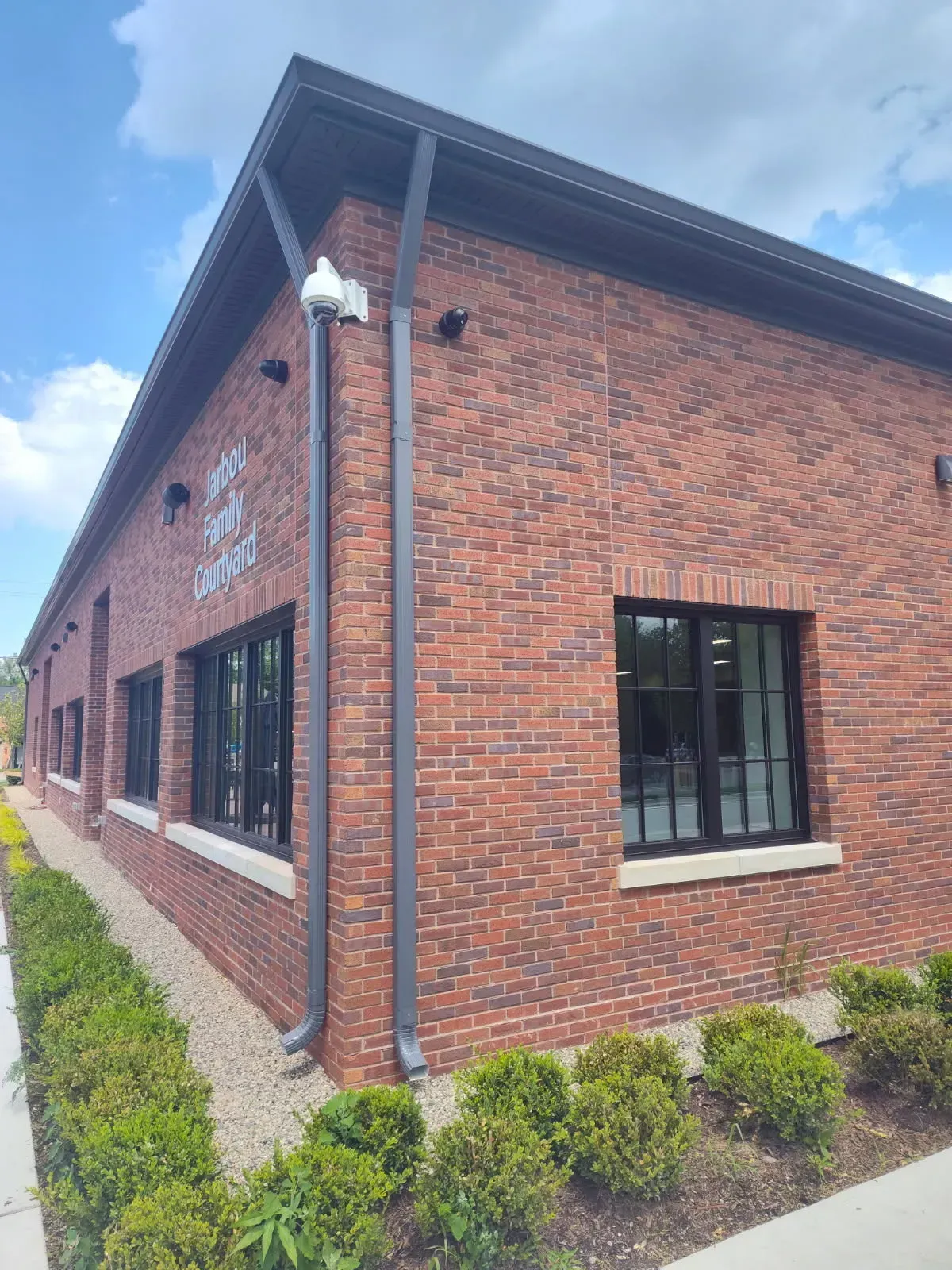 Brick building exterior with black-framed windows, gray downspout, and landscaped shrubs along the wall.