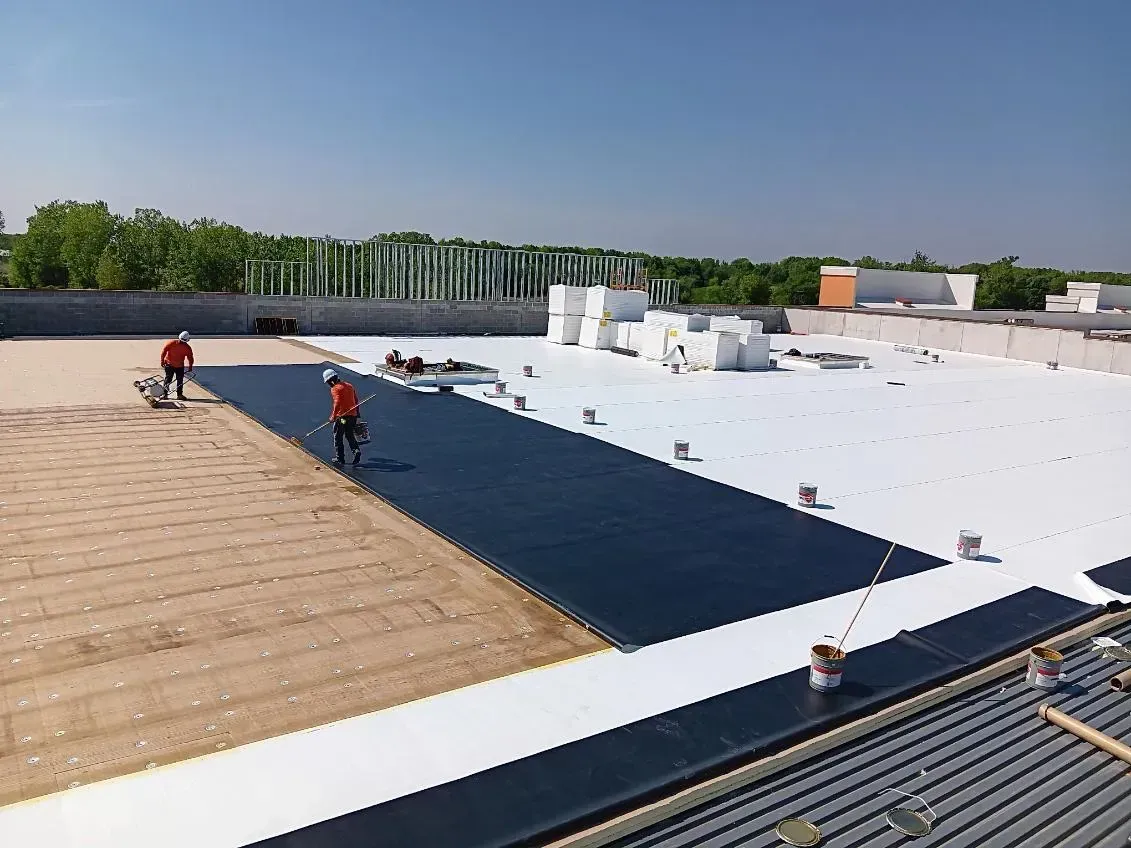 Workers on a flat rooftop installing dark waterproof roofing over a large white roof area.